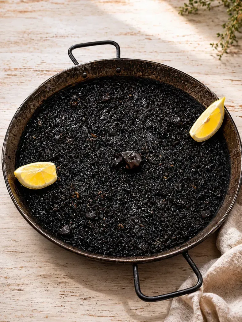 Paellera negra con arroz oscuro, adornada con rodajas de limón sobre una mesa rústica.