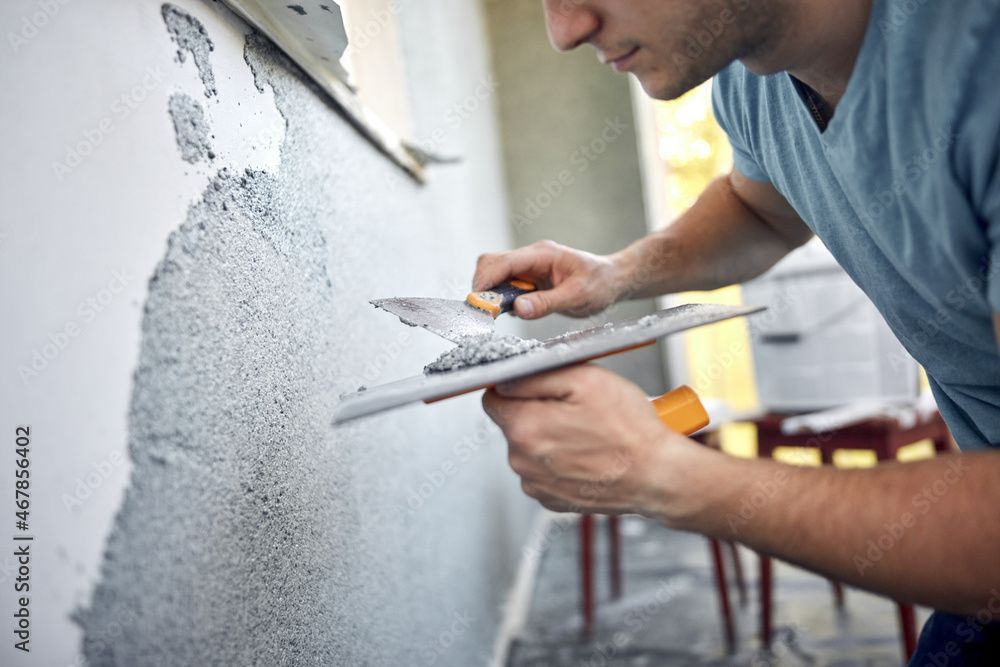Mann trägt mit einer Kelle Putz auf eine Wand auf. Grauer Putz, blaues Hemd, Inneneinrichtung.