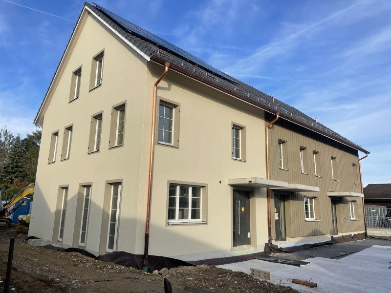 Zweistöckiges Haus mit hellbeigem und grauem Stuck, Kupferdachrinnen, Solarmodulen und einem klaren blauen Himmel.