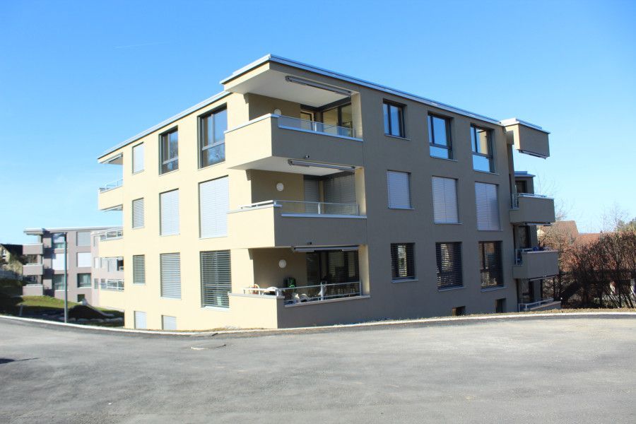 Haus mit rotem Dach, grünem Rasen, Terrasse mit Tisch und Stühlen und umgebender Landschaft an einem bewölkten Tag.