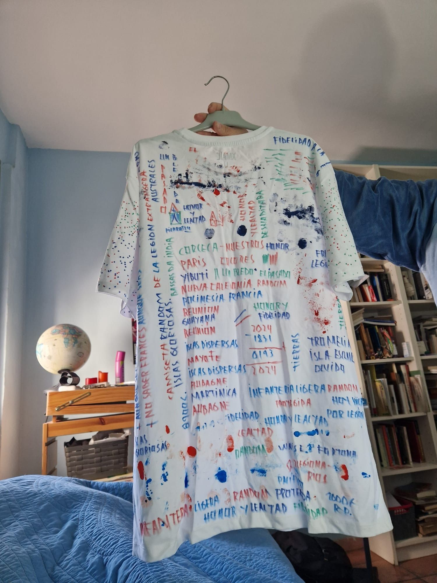 Camiseta blanca cubierta de texto escrito a mano de colores y salpicaduras de pintura, colgada frente a una habitación.