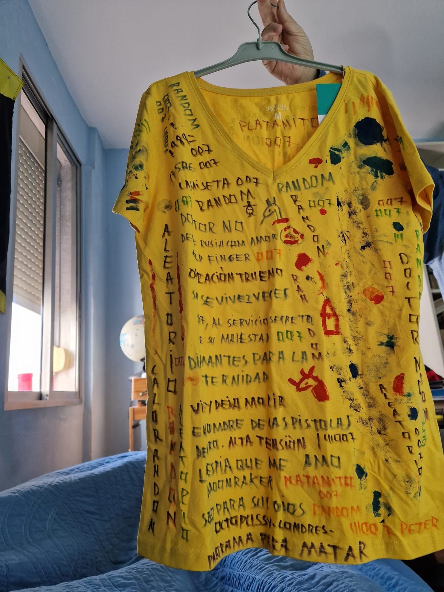 Camiseta amarilla con texto escrito a mano en varios colores, colgada en una habitación.