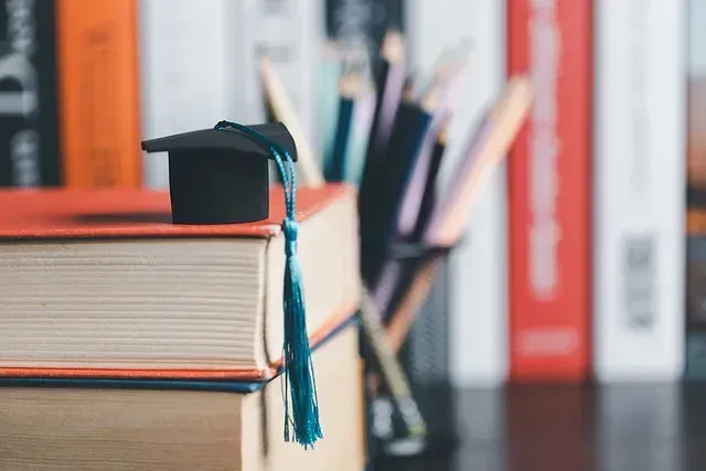 Gorro de graduación con borla azul encima de libros, lápices y libros en el fondo.