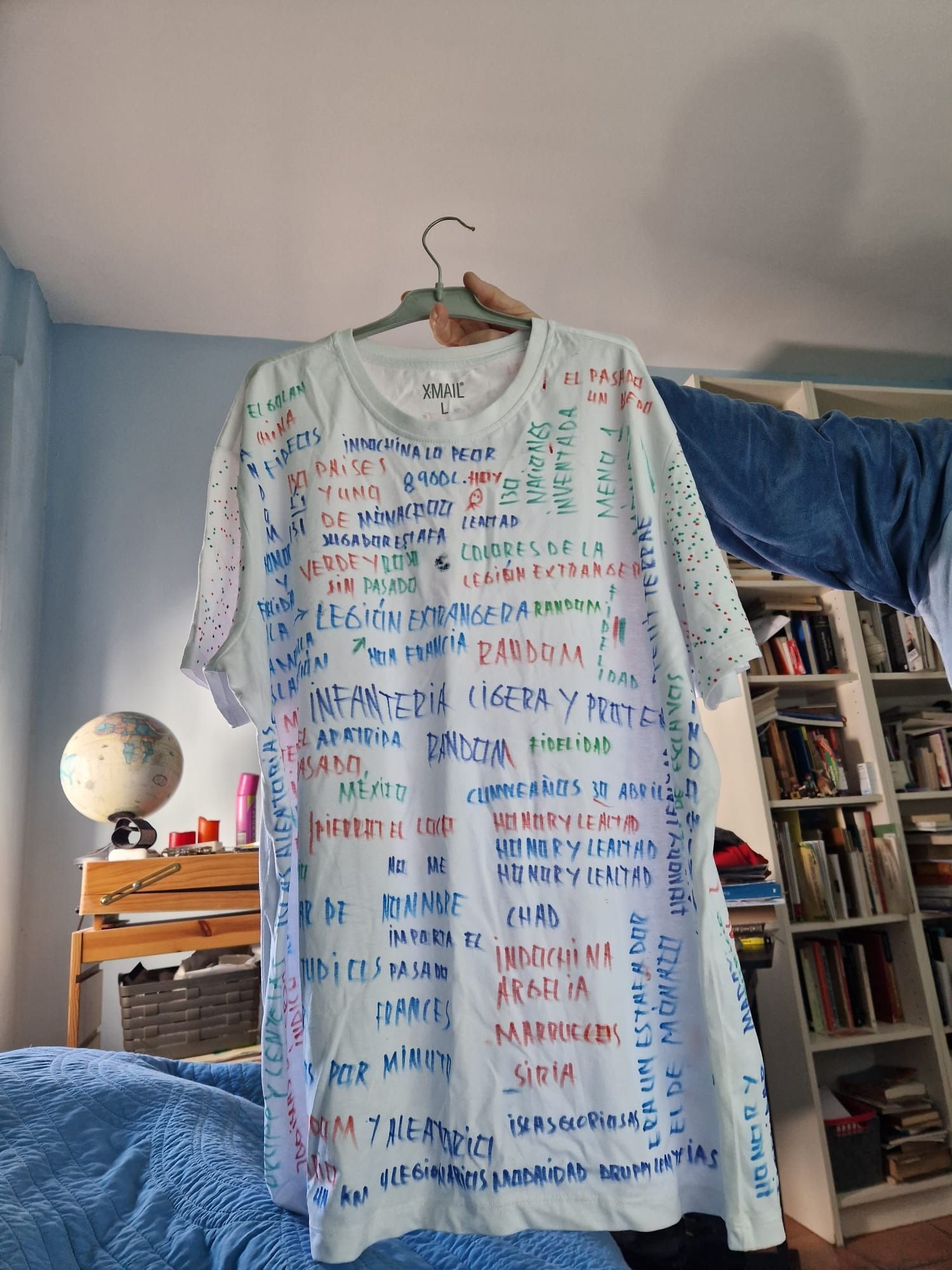 Camiseta blanca con texto escrito a mano en color colgando de una percha de madera en una habitación con una estantería y un globo terráqueo.