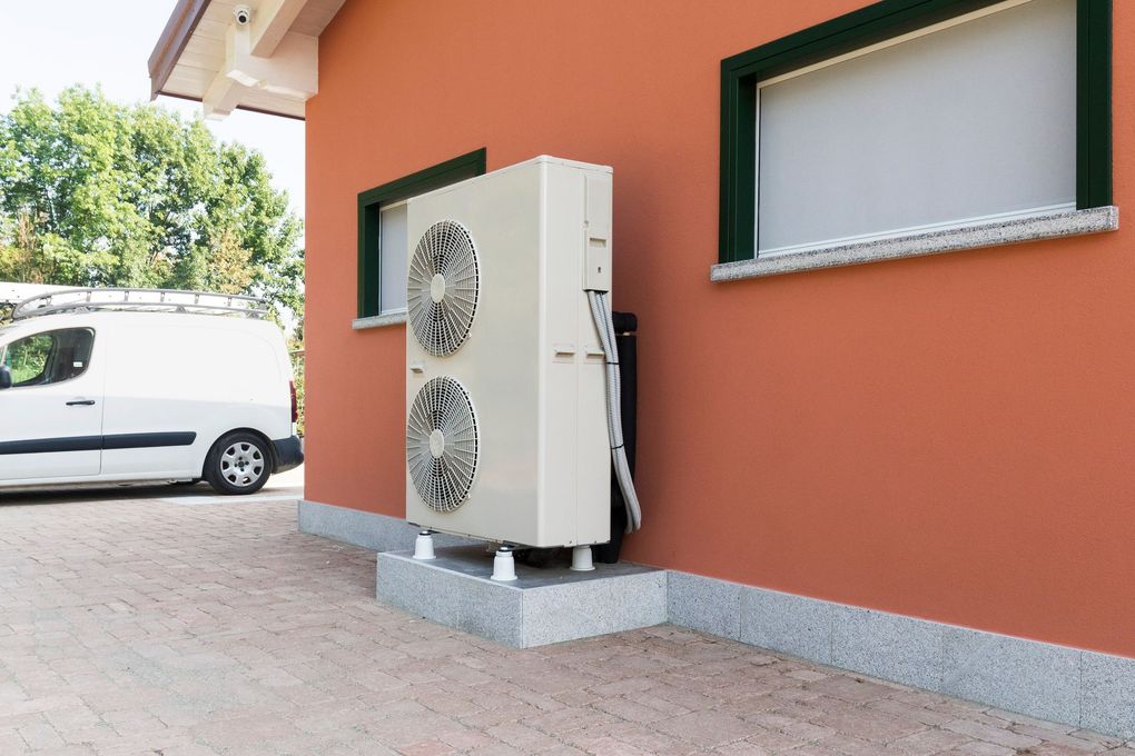Unité de climatisation extérieure à côté du mur d'une maison en terre cuite, avec une camionnette blanche garée à proximité.
