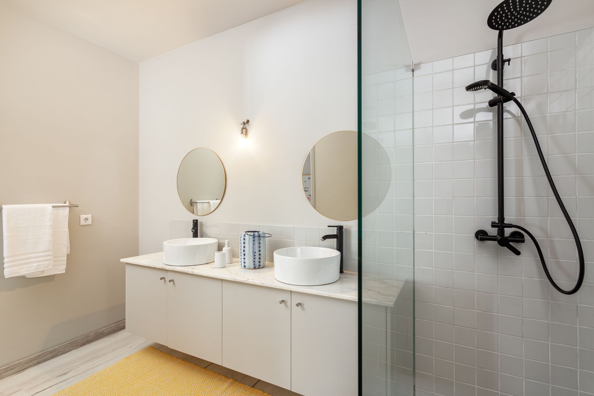 Salle de bains lumineuse avec double vasque, miroirs ronds et douche en verre avec robinetterie noire.