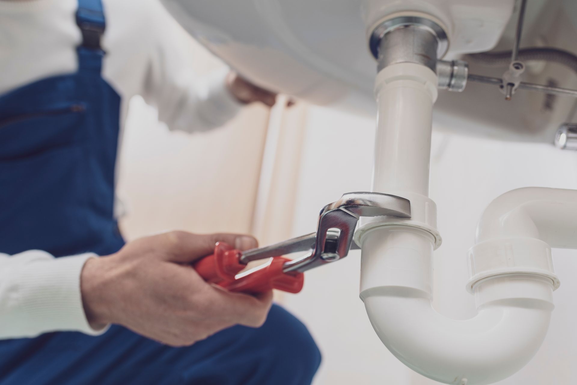 Un plombier utilise une clé rouge pour resserrer le tuyau d'évacuation blanc sous un lavabo.