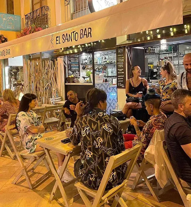 Un grupo de personas están sentadas en mesas bajo un cartel que dice el santo bar.
