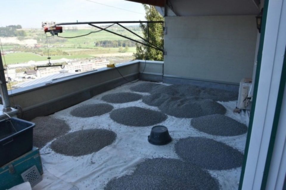 Eine Dachkonstruktion mit Kieskreisen auf weissem Untergrund. Ein Eimer und eine Werkzeugkiste stehen daneben.