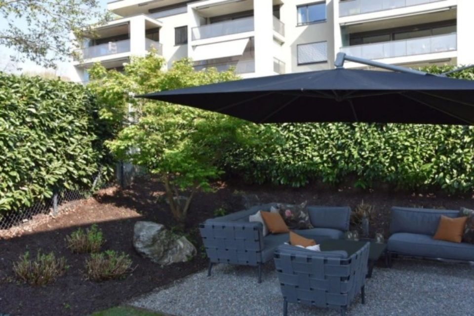 Terrasse im Freien mit schwarzem Sonnenschirm, grauen Sitzgelegenheiten und üppiger grüner Hecke.