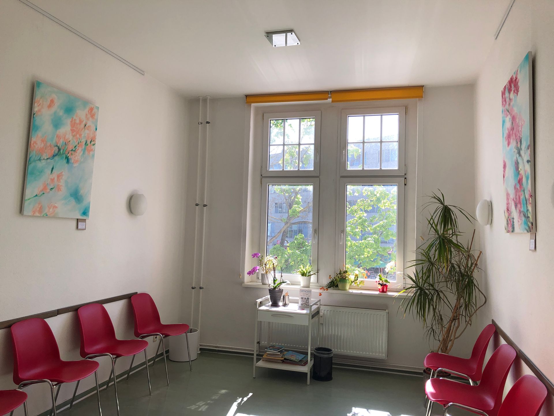 Heller Warteraum mit roten Stühlen an den Wänden, zwei bunten Gemälden mit Blütenmotiven und großen Fenstern, durch die Tageslicht einfällt. Auf der Fensterbank stehen Pflanzen, daneben eine Palme im Topf.
