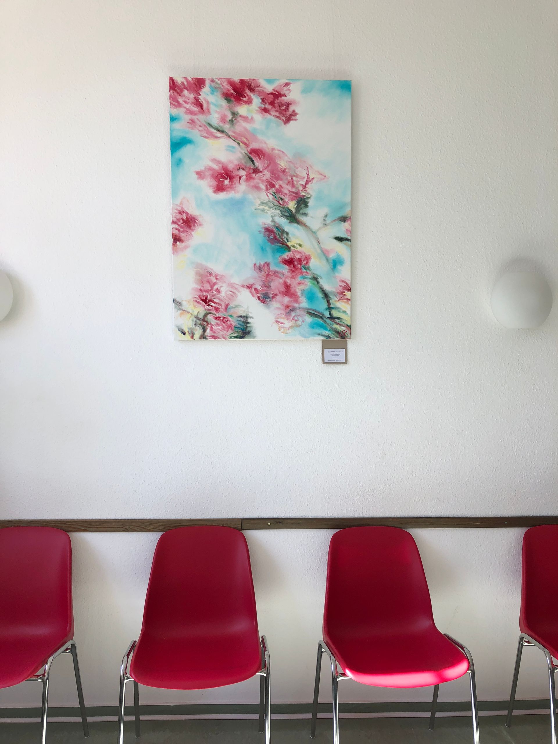 Ein heller Warteraum mit drei roten Stühlen vor einer weißen Wand. Darüber hängt ein farbenfrohes Gemälde mit rosa Blüten vor blau-weißem Hintergrund.