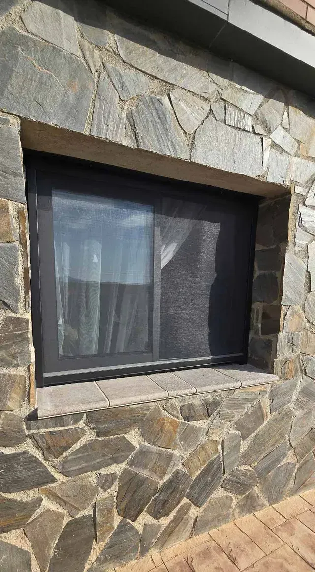 Muro exterior de piedra con una ventana de marco oscuro y un camino de ladrillos debajo.