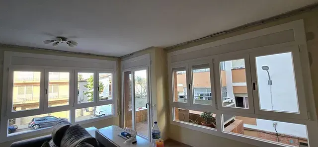 Sala de estar luminosa con grandes ventanales, molduras blancas y vistas a los edificios vecinos.