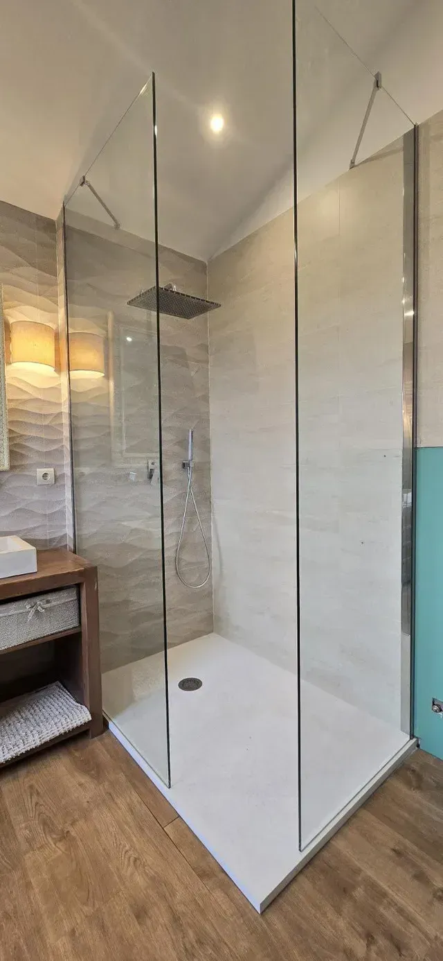 Baño moderno con ducha acristalada, paredes de azulejos blancos y suelo con aspecto de madera.