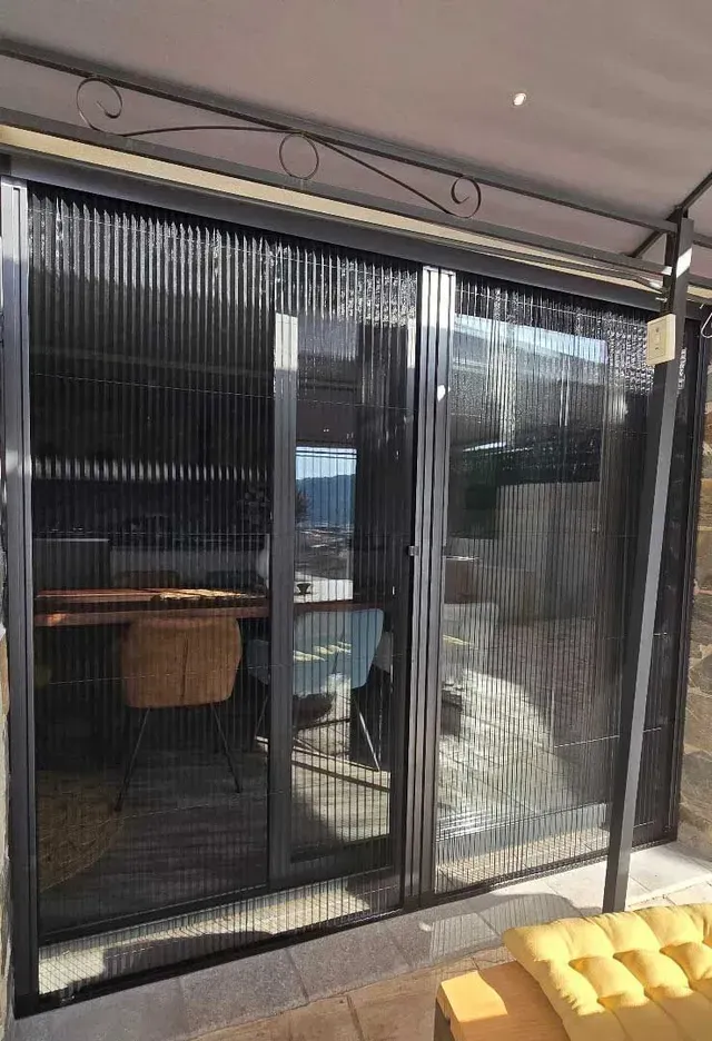 Puertas corredizas de malla negra en un balcón con asientos al aire libre visibles desde el interior.