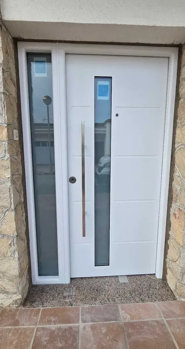 Entrada principal con puerta blanca, panel lateral de cristal estrecho y umbral de baldosas marrones.