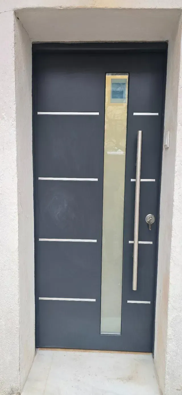 Puerta de entrada moderna de color gris con panel de vidrio vertical y manija larga de metal en un marco de pared blanco.