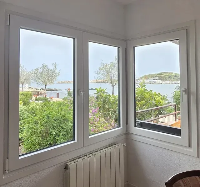 Ventana esquinera con marcos blancos con vistas a un jardín verde y a aguas tranquilas.