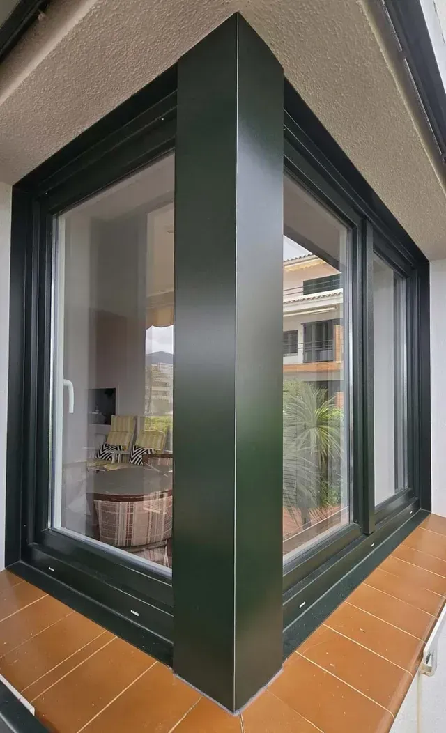 Rincón de una casa moderna con marcos de ventanas de color verde oscuro y una terraza de madera con vistas al interior.