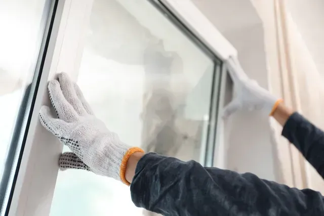 Manos enguantadas limpian una ventana con marco blanco con un paño.