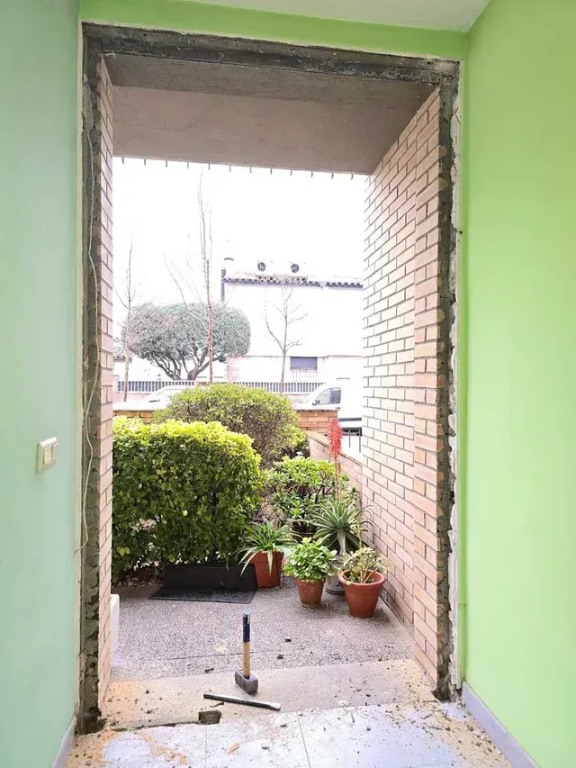 Entrada abierta a un pequeño patio ajardinado con macetas y una pared de ladrillos.