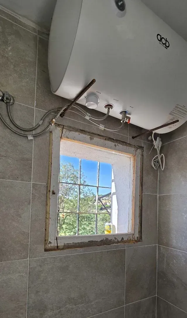 Pequeño baño en construcción con azulejos y una ventana que da a árboles y cielo azul.