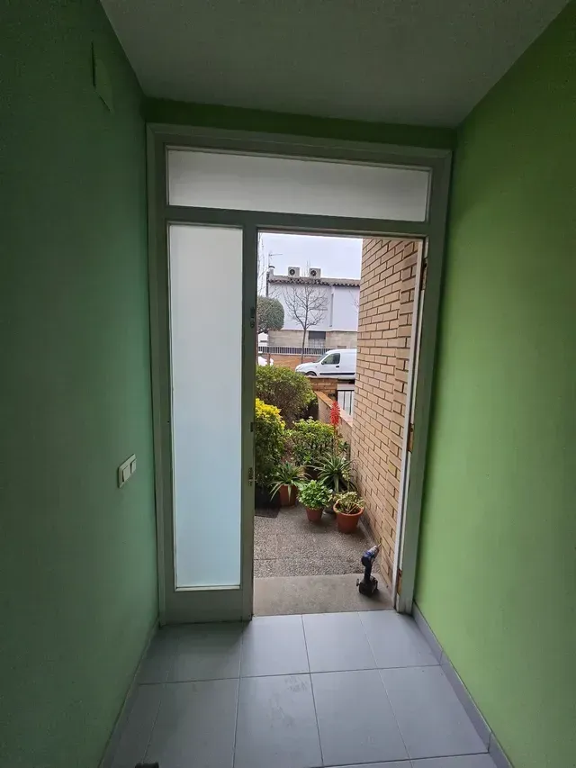 Un pasillo verde conduce a una puerta principal abierta, con un pequeño gato sentado afuera, junto a la entrada.