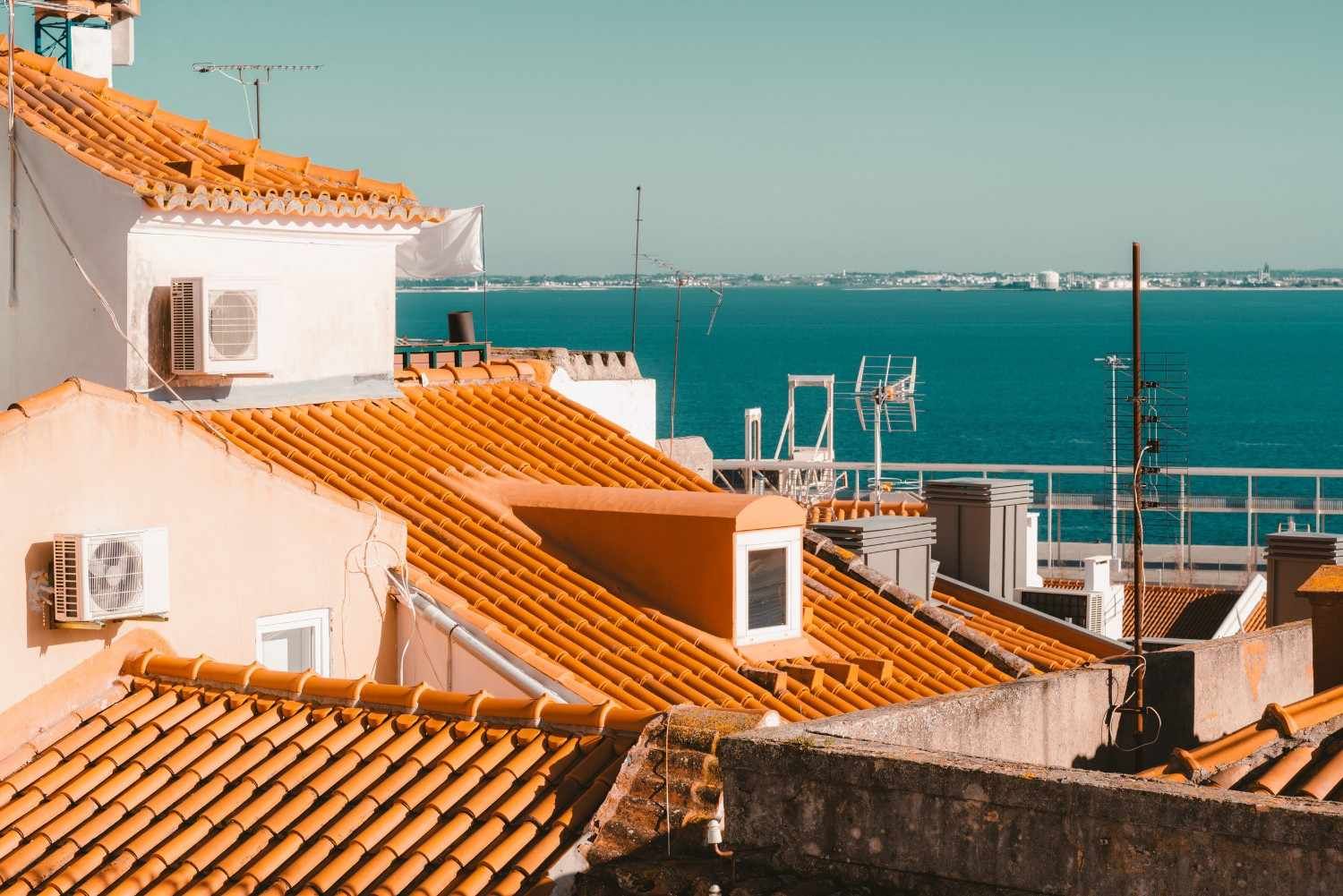 Tejados blancos y de terracota con vistas a un mar turquesa y un puerto lejano bajo un cielo despejado.