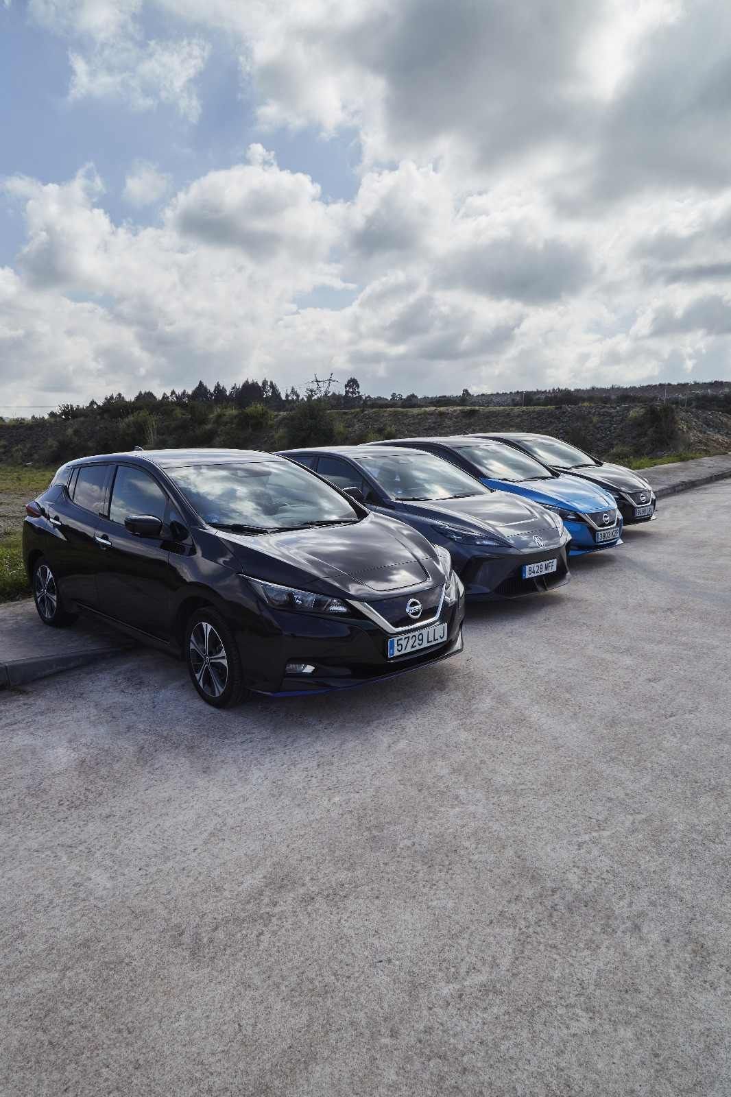 Cinco coches eléctricos aparcados en fila sobre el asfalto con fondo de cielo nublado.