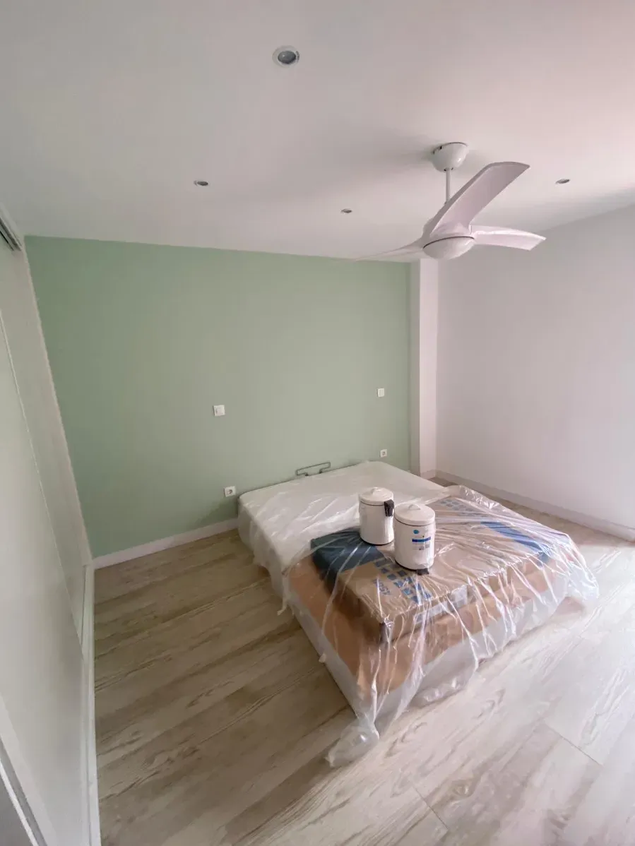 Dormitorio minimalista con cama, pared decorativa de color verde pálido, ventilador de techo blanco y suelo de madera clara.