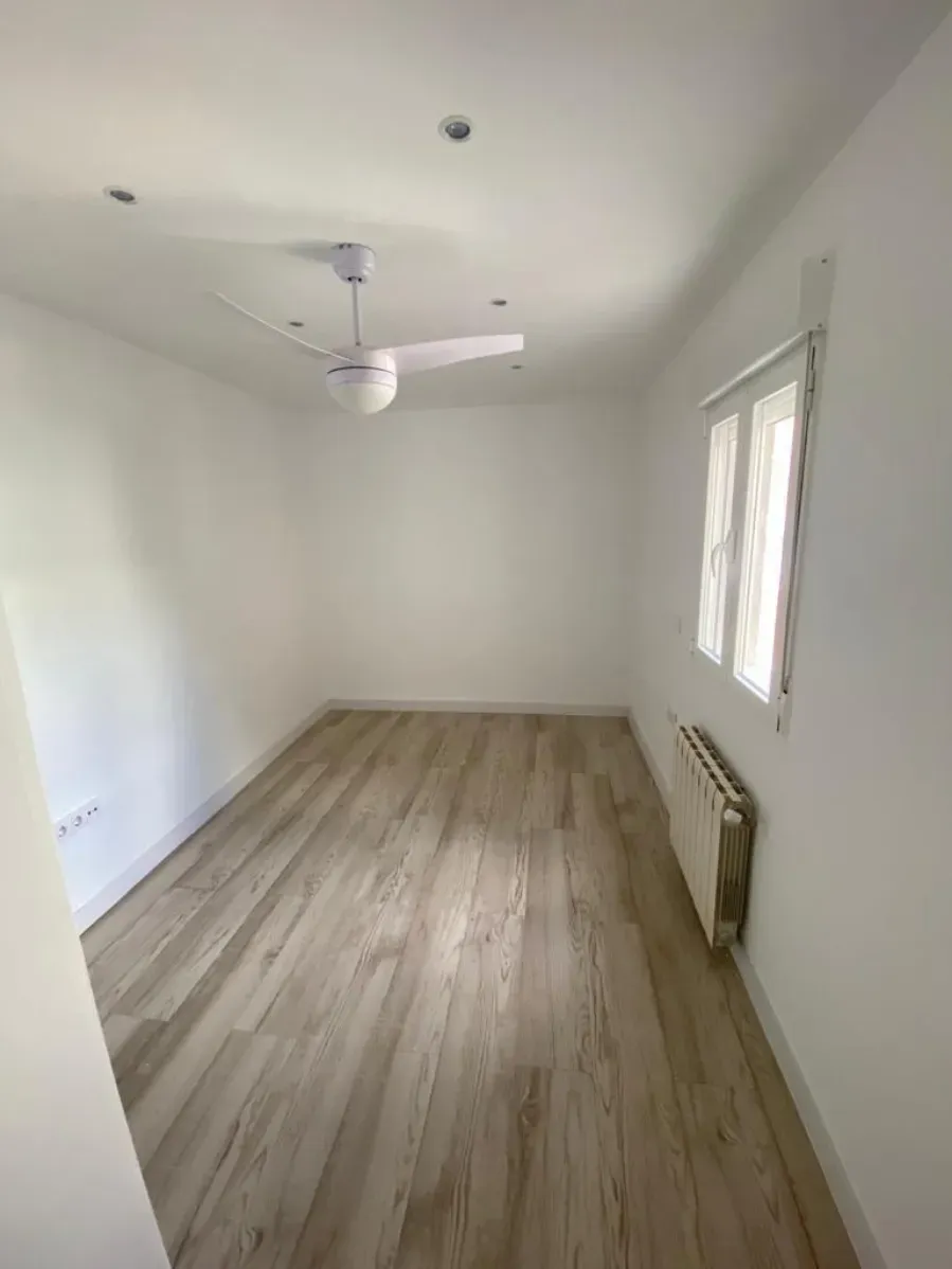 Habitación blanca vacía con suelo de imitación madera, ventilador de techo y ventanas altas a la derecha.
