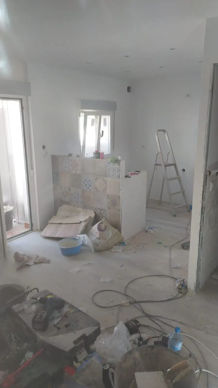 Habitación en obras con escombros, escalera, materiales de pintura y una pared parcialmente alicatada.