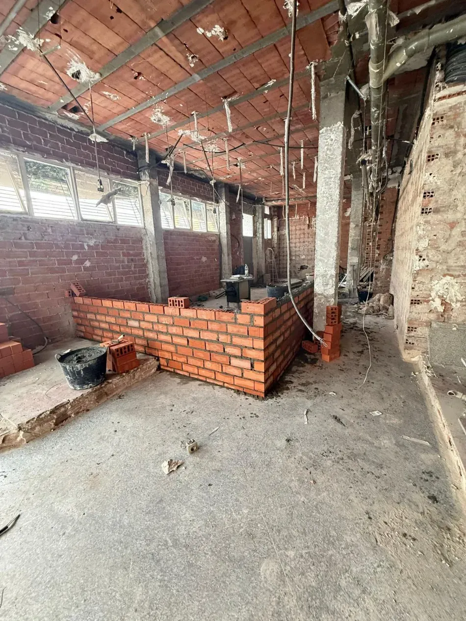 Interior de ladrillo sin terminar con piso de concreto, vigas a la vista y un tabique bajo de ladrillo en construcción.