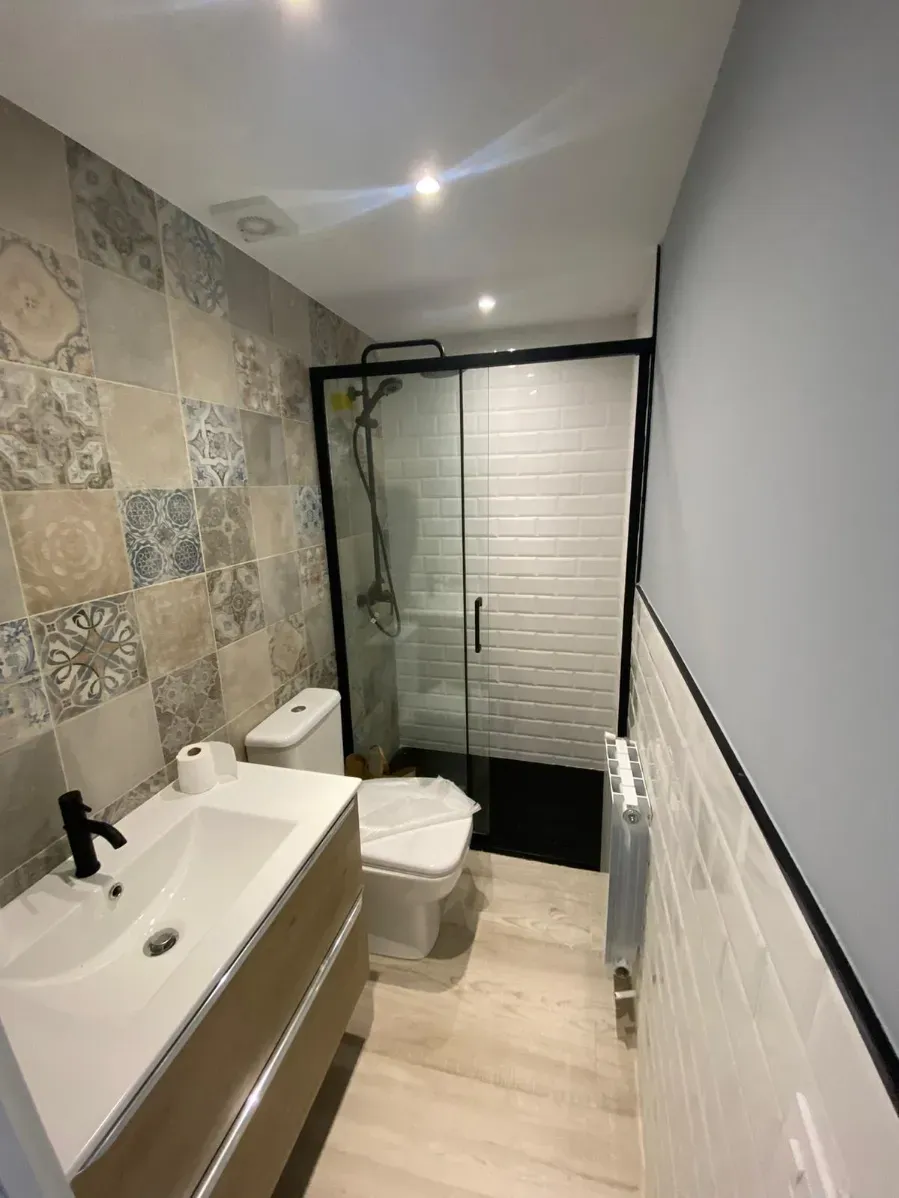 Baño moderno y estrecho con lavabo, inodoro y ducha de cristal, con azulejos de pared con estampado de piedra y suelo de madera clara.