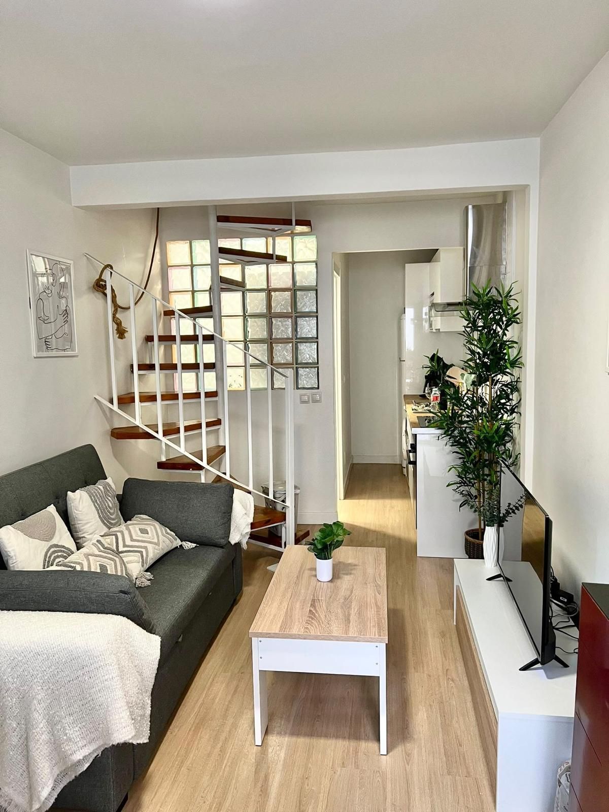 Acogedora sala de estar con sofá modular beige, televisor, alfombra turquesa, mesa de centro, estantes y planta colgante.