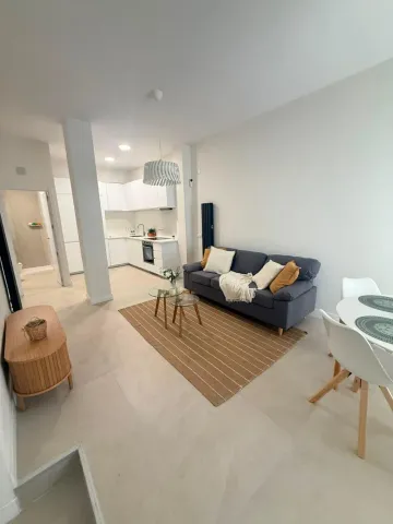 Sala de estar luminosa y minimalista con sofá gris, alfombra tejida, mesa de comedor blanca y cocina abierta.