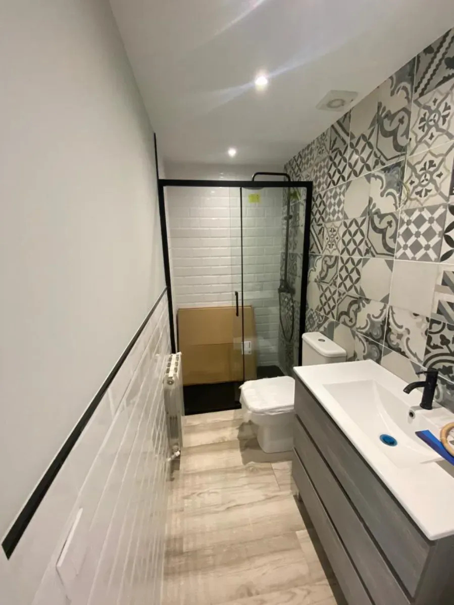 Baño estrecho con pared de azulejos estampados, lavabo blanco, ducha de cristal y un inodoro cerca del fondo.