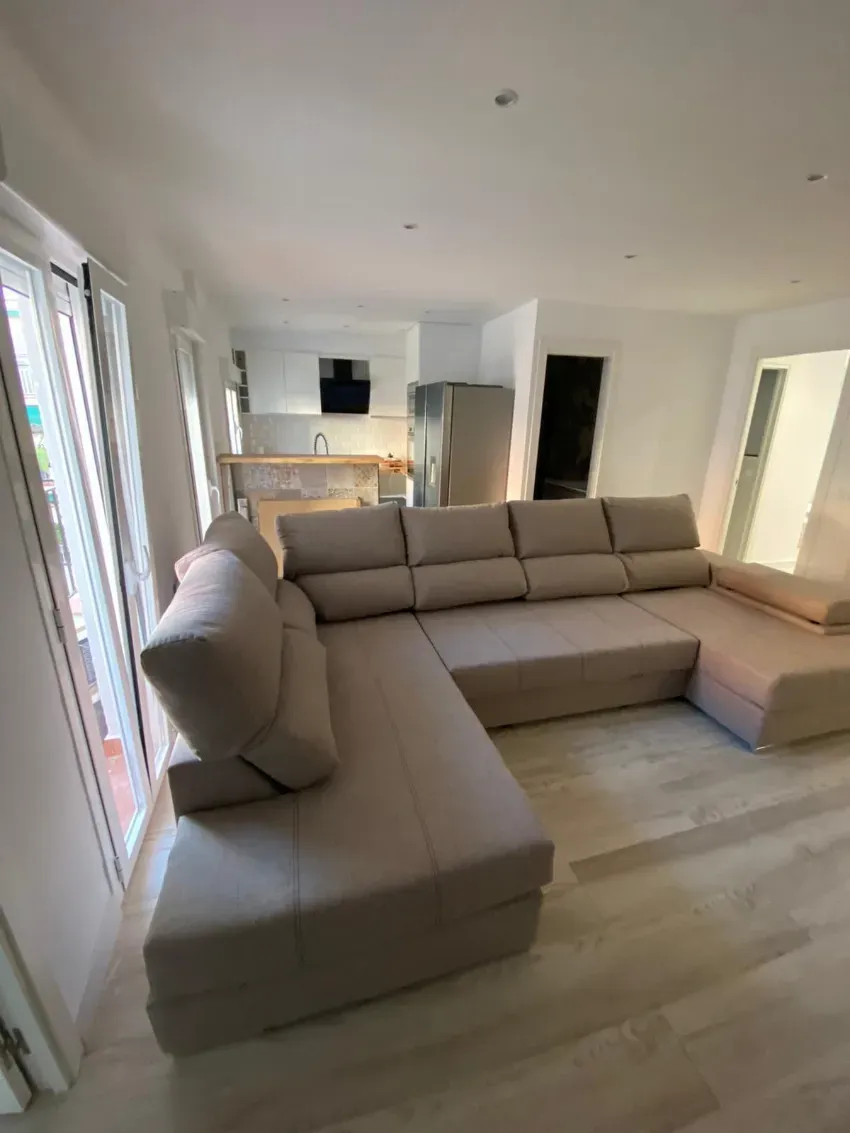 Sala de estar moderna con sofá seccional beige, cocina abierta y grandes ventanales con cortinas.