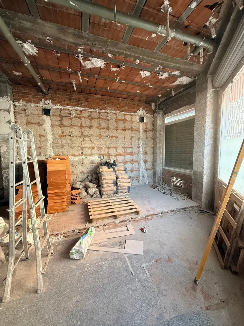 Habitación abandonada en obras con paredes de ladrillo visto, andamios, escalera y materiales apilados en el suelo.