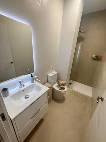 Baño moderno con lavabo blanco, espejo, inodoro y ducha a ras de suelo con azulejos beige.