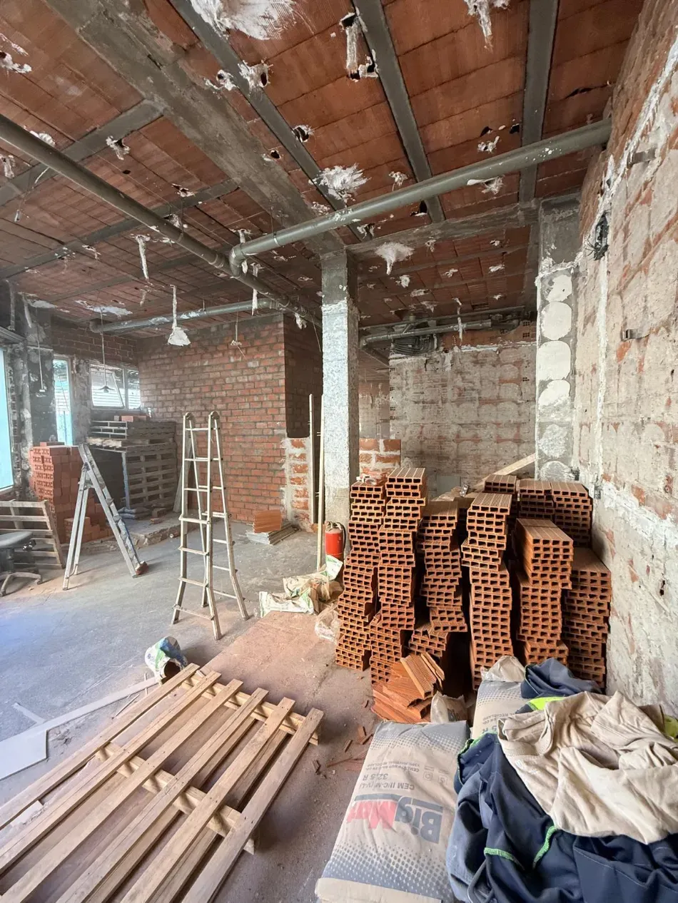 Interior de una obra en construcción con paredes de ladrillo visto, ladrillos apilados, escaleras y herramientas sobre un suelo polvoriento.
