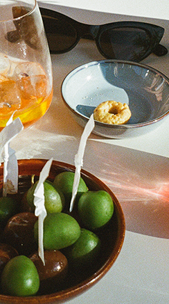 Auf einem Tisch steht eine Schale mit grünen Oliven neben einem Glas Wein und einer Sonnenbrille.
