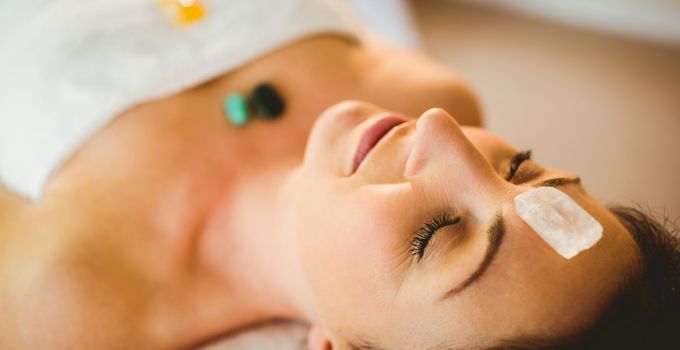 Femme recevant un soin du visage, des pierres sur le visage et le torse, les yeux fermés, allongée sur une table de massage.