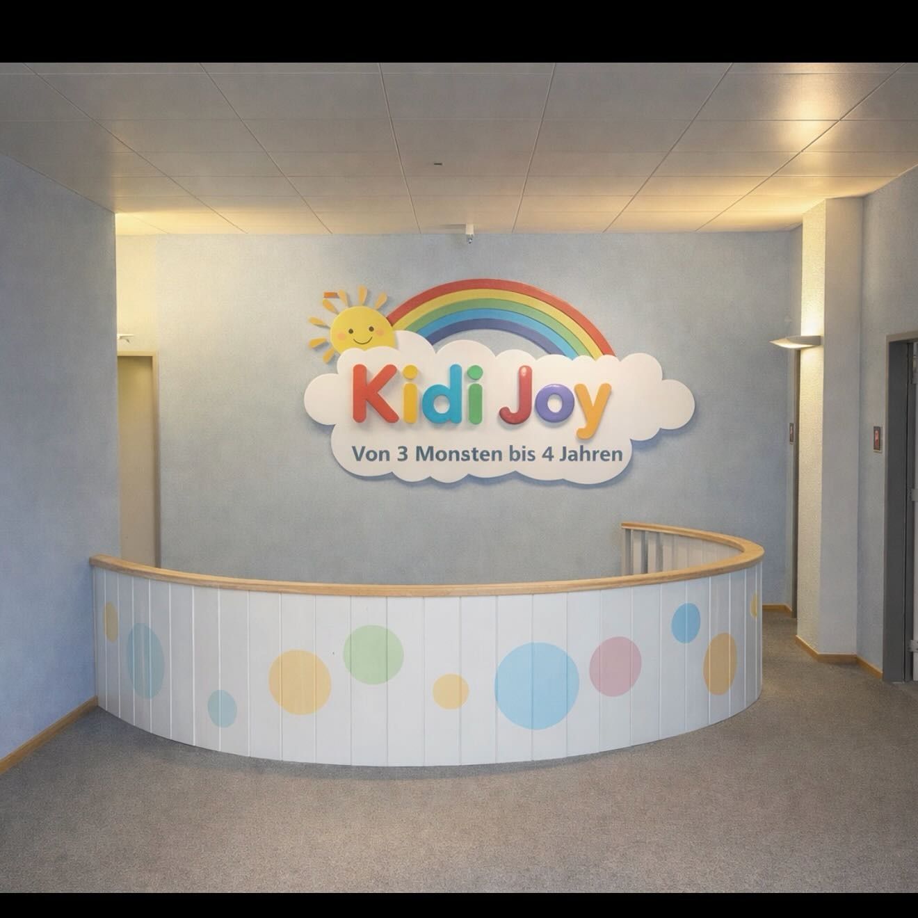 Empfangstresen der Kindertagesstätte Kidi Joy mit einem farbenfrohen Logo aus Regenbogen, Sonne und Wolke auf hellblauer Wand.