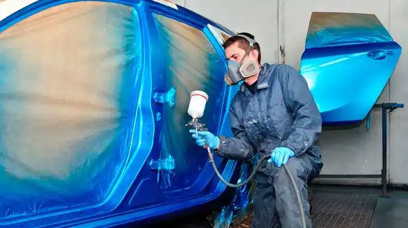 Un hombre que lleva una máscara está pintando con aerosol un coche azul.