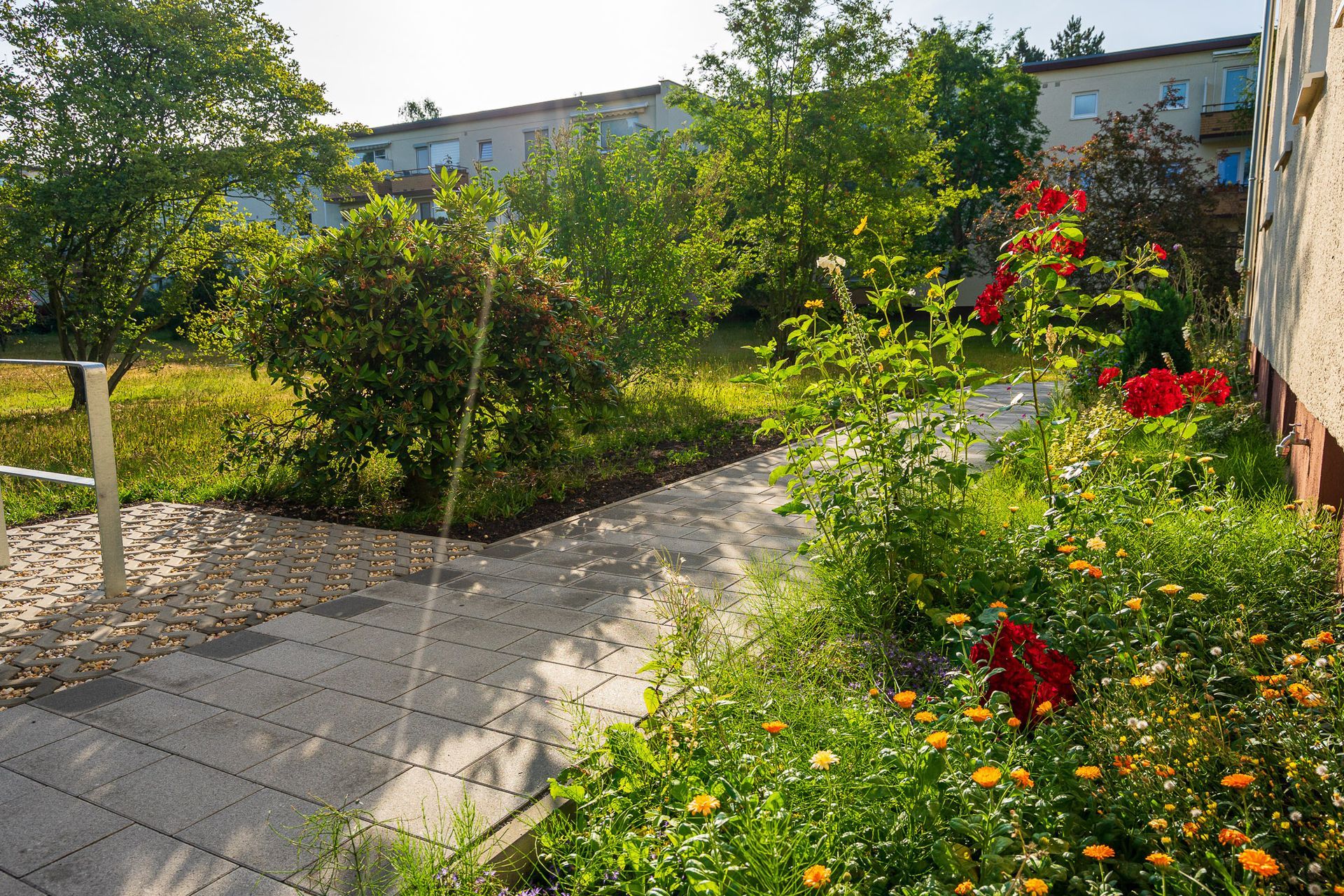 Gepflasterter Weg mit grauen Steinen in moderner Wohnsiedlung – umgeben von gepflegten Grünflächen und dekorativen Pflanzen. Die harmonische Wegführung verbindet Eingangsbereiche und schafft eine naturnahe Atmosphäre. Ideal für nachhaltige Außenanlagen, Vorgartengestaltung und urbane Wohnkonzepte.
