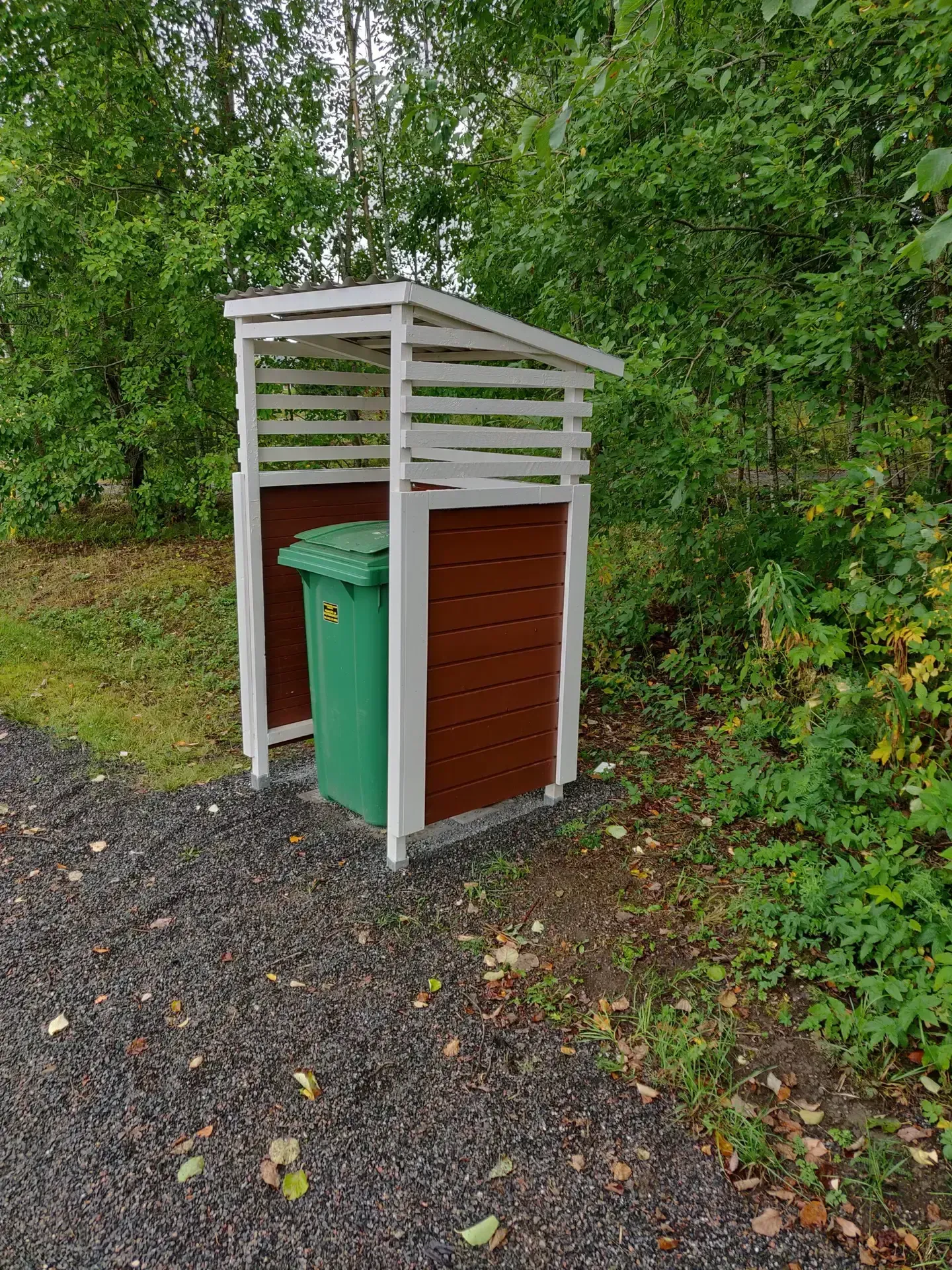 Vihreä roskakori on ulkona pienen, katetun puurakenteen sisällä, jossa on valkoiset kehykset ja punertavanruskeat seinät.