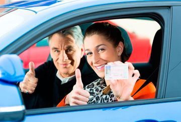 Una mujer está sentada en un automóvil sosteniendo una licencia de conducir y haciendo un gesto de aprobación con el pulgar hacia arriba.
