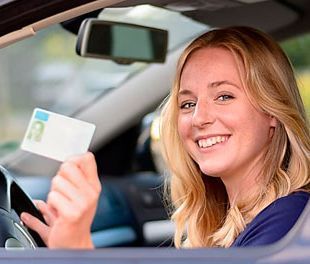 Una mujer está sentada en un automóvil sosteniendo un permiso para conducir y sonriendo.
